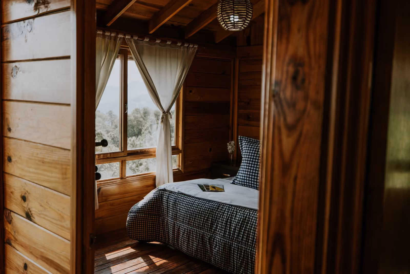 Une petite chambre lambrissée de bois, literie à carreaux et fenêtre à rideaux ouvrant sur des collines boisées.