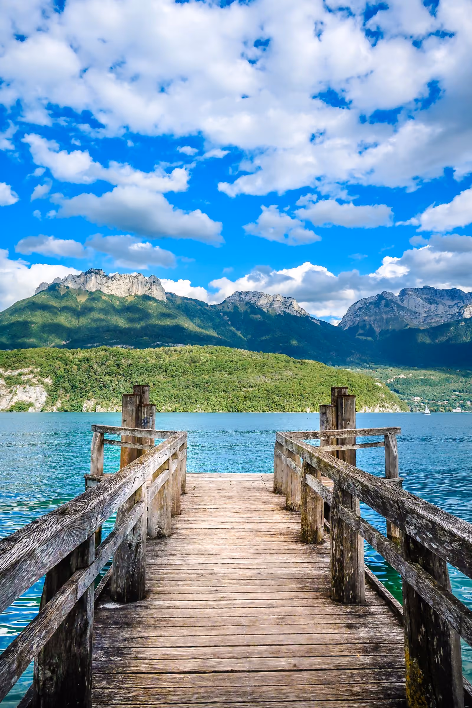 Un ponton de bois patiné s’avançant dans l’eau turquoise du lac d’Annecy, la crête calcaire des Dents de Lanfon en arrière-plan.