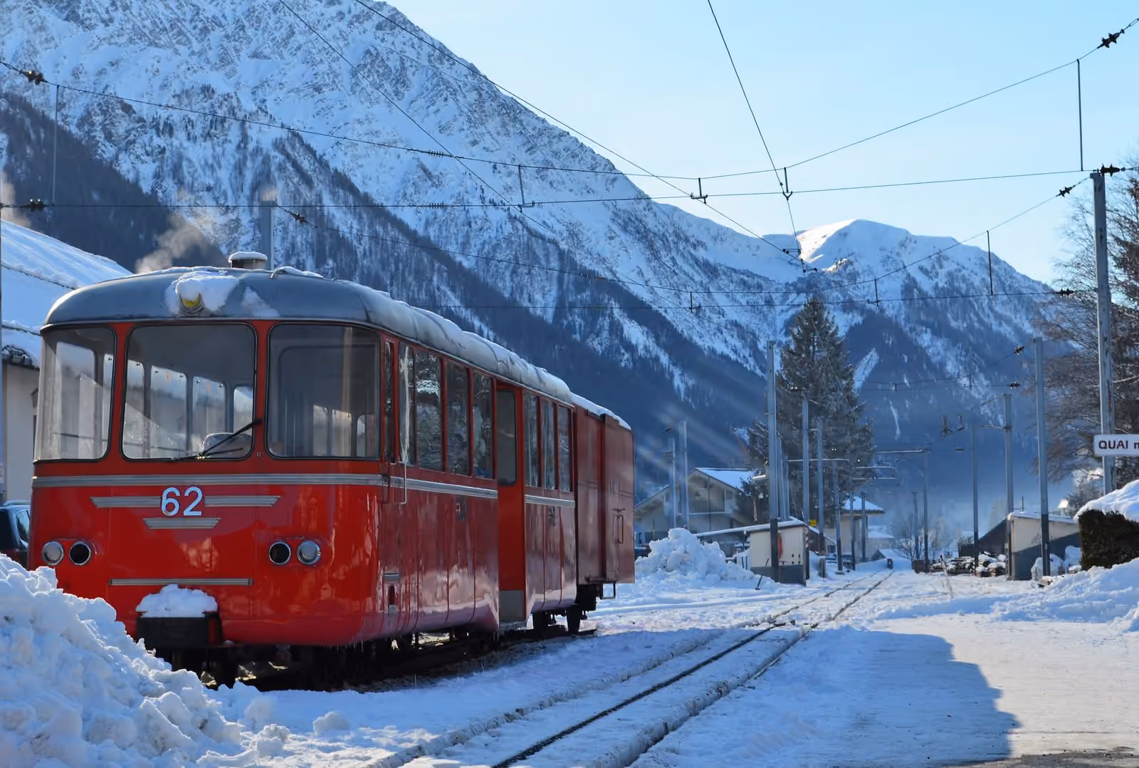 Un autorail rouge à voie métrique à quai sous la neige, au pied de versants abrupts.