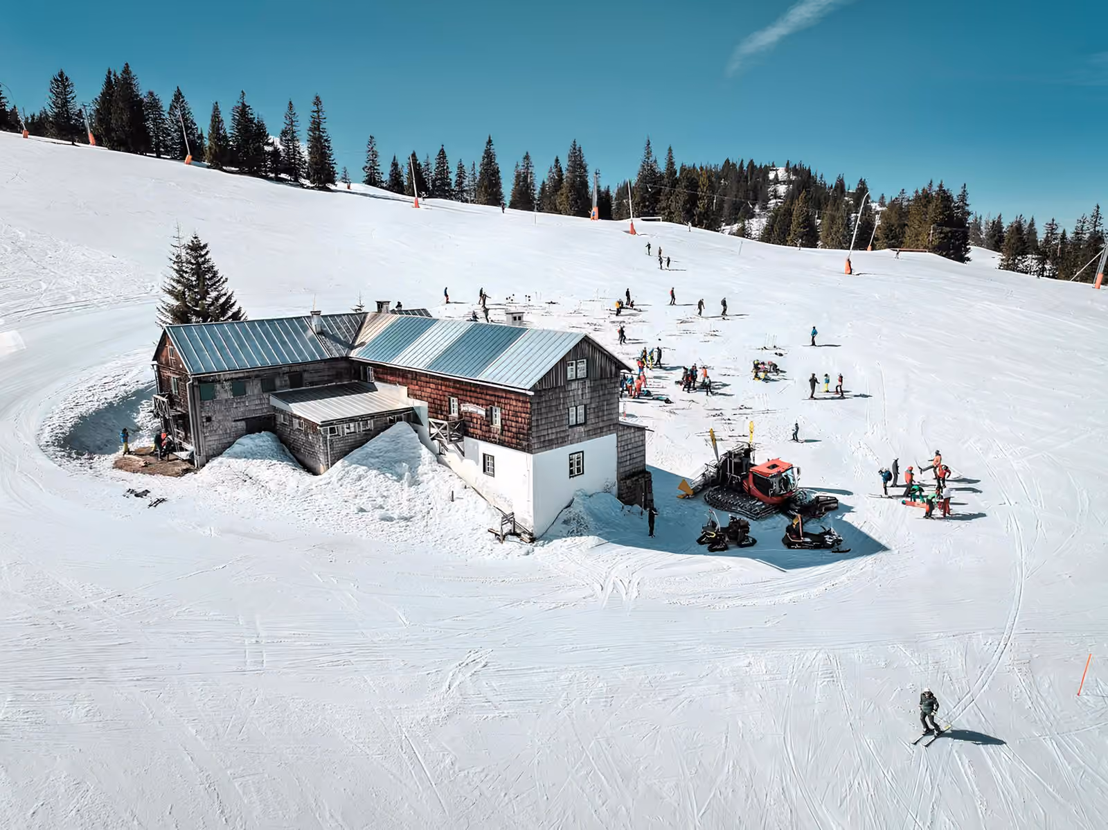 Un chalet-hôtel posé directement sur une piste damée, skieurs et groupe d’école de ski rassemblés devant sa porte.